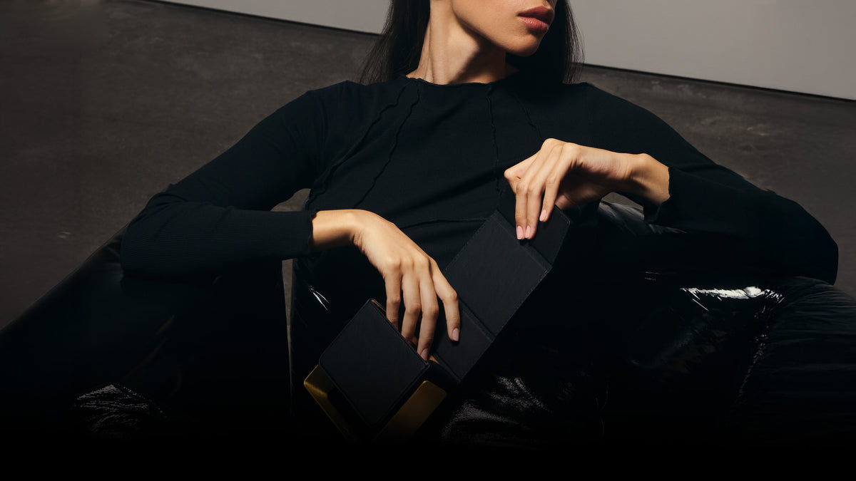 Woman in a black top reclining in a glossy black chair, opening a black bio-leather BEAM handbag with a brushed copper frame
