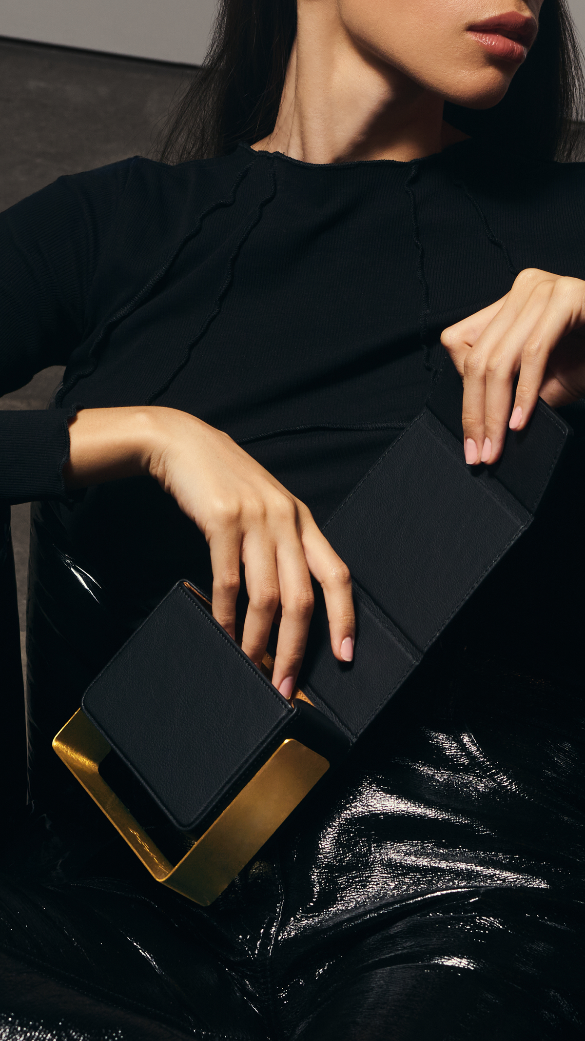 Woman in a black top reclining in a glossy black chair, opening a black bio-leather BEAM handbag with a brushed copper frame
