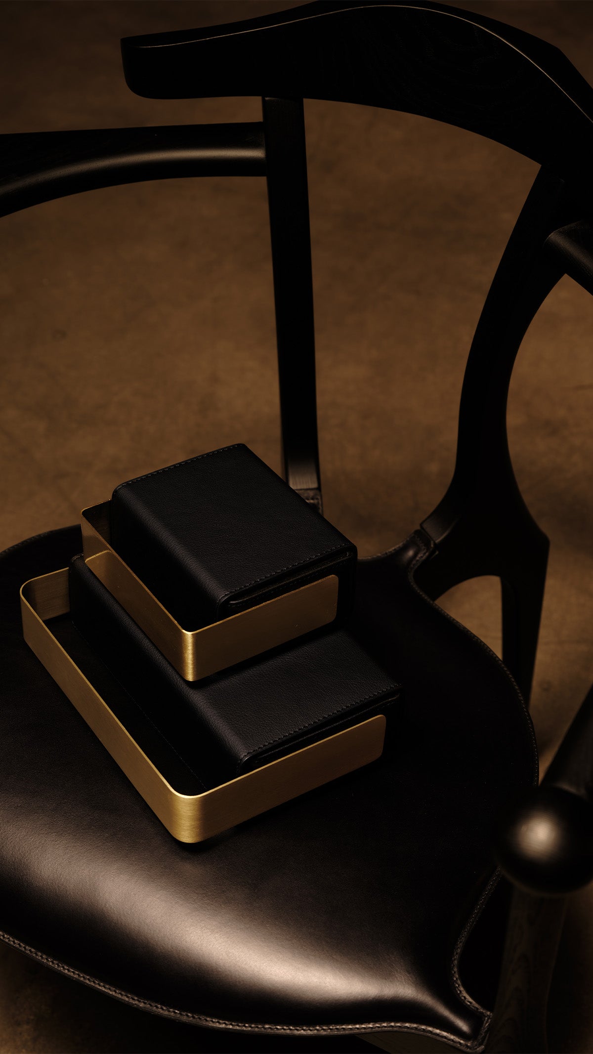 Two black bio-leather BEAM vessels with brushed copper frames stacked on the seat of a sculptural black wooden chair